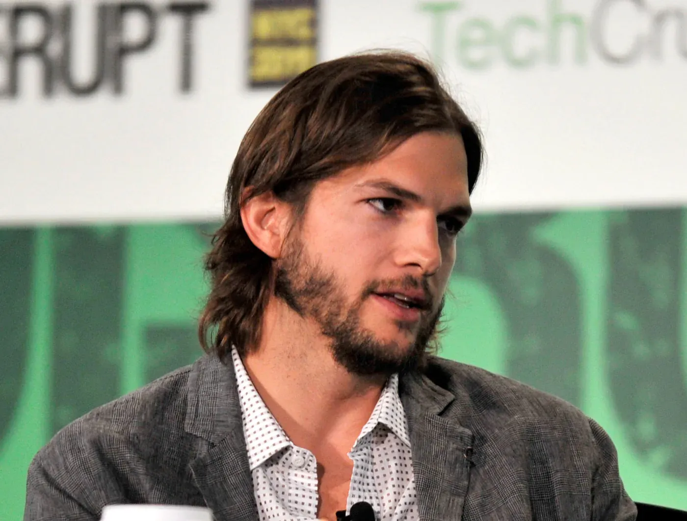 Ashton Kutcher seated on a conference panel in a grey blazer, shoulder-length hair and a beard, turned in three-quarter profile.