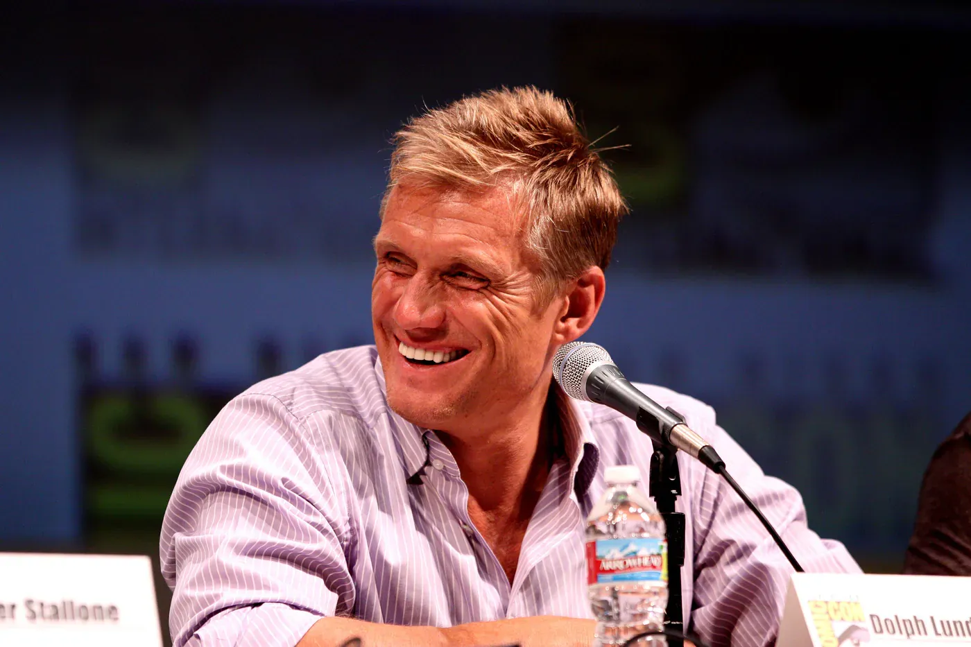 Dolph Lundgren laughing at a panel table behind a microphone, wearing a pink striped shirt.