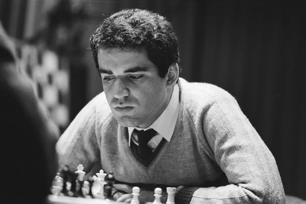 Garry Kasparov leaning over a chessboard in a light sweater and tie, studying the position. Black and white.