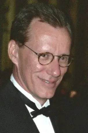 James Woods in a tuxedo and bow tie with round glasses, smiling toward the camera.