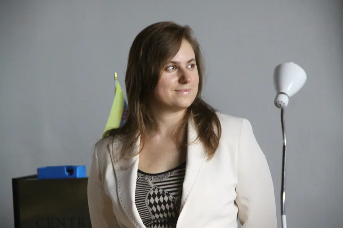 Judit Polgár in a white blazer, seated and looking off to one side against a plain grey background.