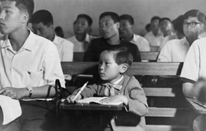 Kim Ung-yong as a young child at a lecture-hall desk with a pencil and open notebook, older students seated behind him. Black and white.
