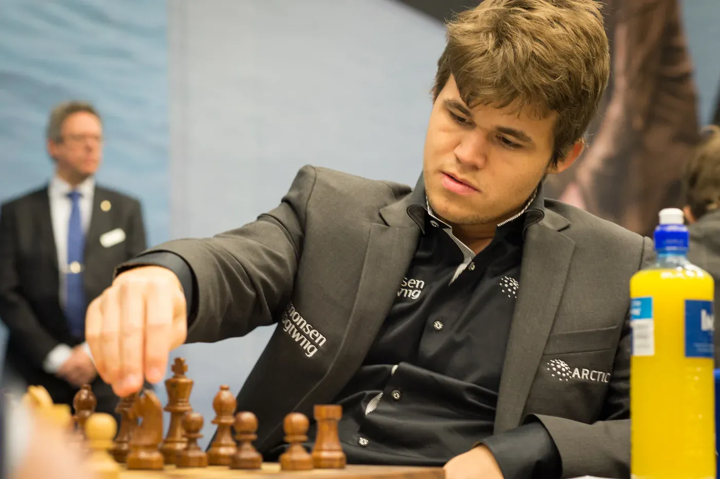 Magnus Carlsen reaching to move a piece at the chessboard, in a dark shirt and grey jacket.