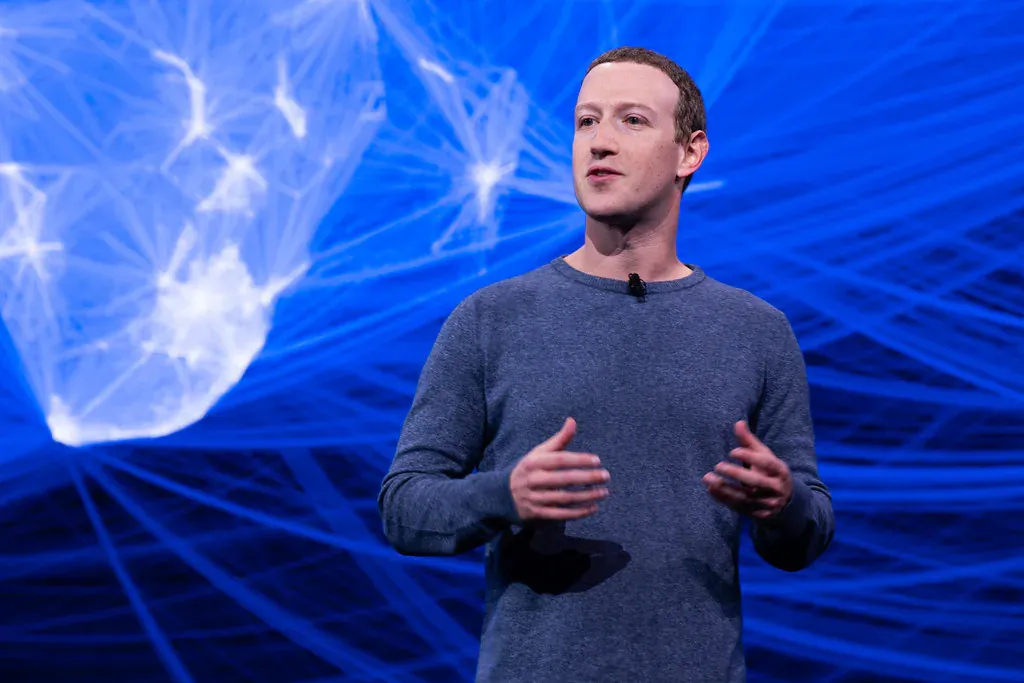 Mark Zuckerberg speaking on stage in a grey sweater, hands raised mid-gesture, against a blue backdrop.