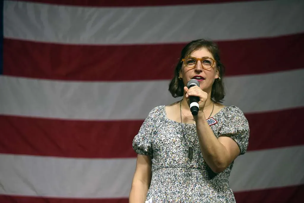 Mayim Bialik speaking into a handheld microphone in a patterned dress, a large American flag behind her.