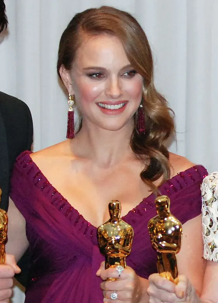 Natalie Portman smiling in a magenta gown with drop earrings, Academy Award statuettes held in front of her.
