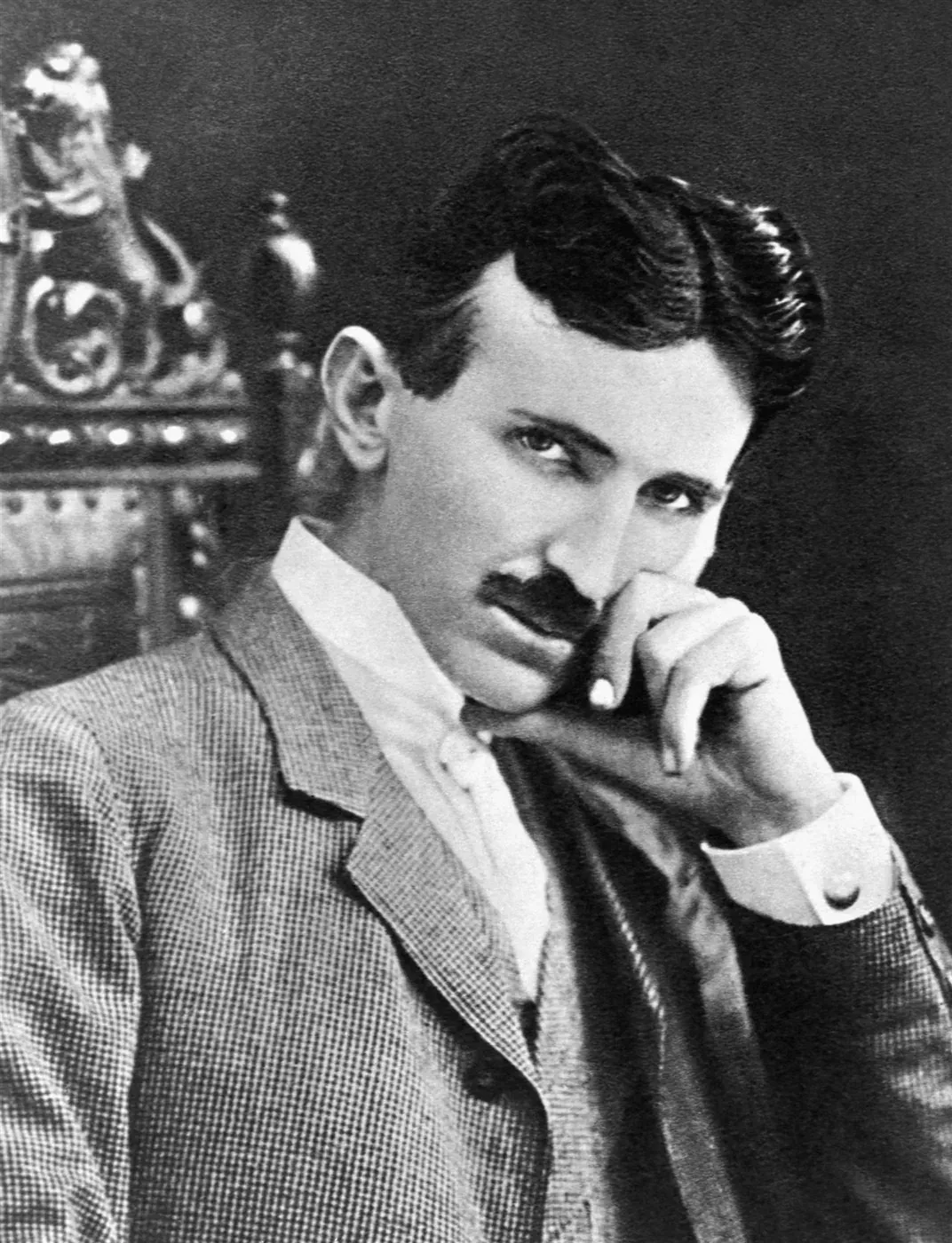 Nikola Tesla seated with a hand resting against his chin, in a suit, beside an ornately carved chair back. Black and white.