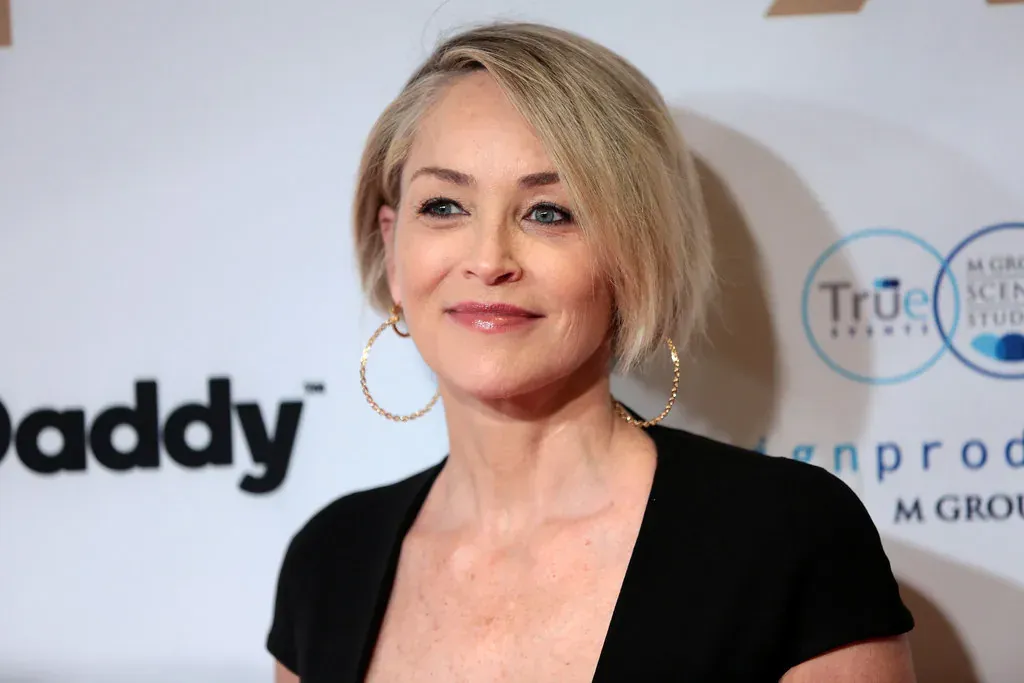 Sharon Stone at a press event with short blonde hair, hoop earrings and a black top, in front of a sponsor backdrop.