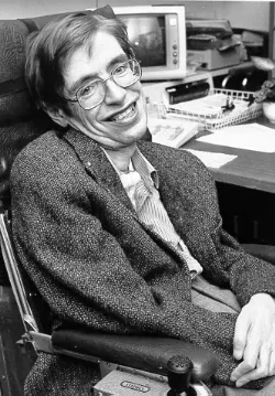 Stephen Hawking smiling in his wheelchair, wearing glasses and a tweed jacket, an office desk behind him. Black and white.