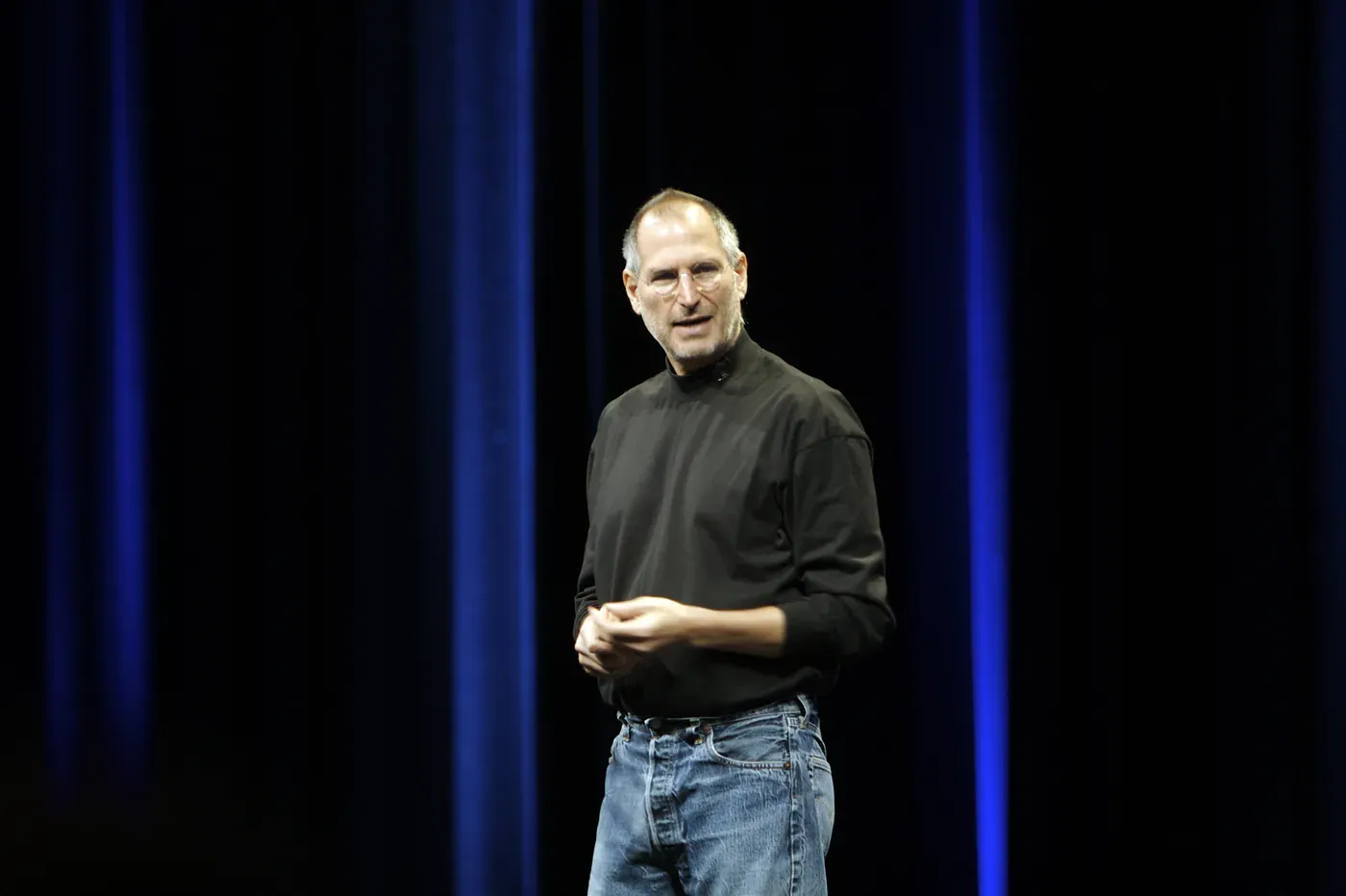 Steve Jobs on a darkened stage in his black turtleneck and jeans, a blue curtain behind him.