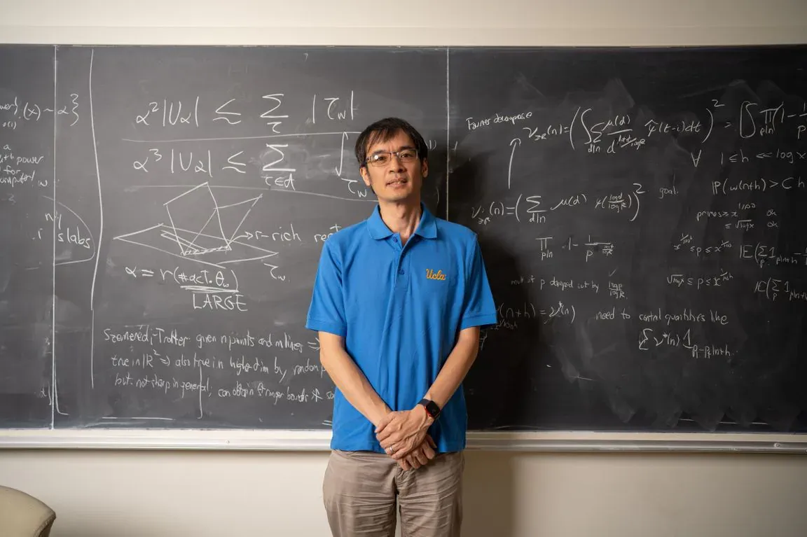 Terence Tao standing in front of a blackboard covered in handwritten equations, in a blue polo shirt.
